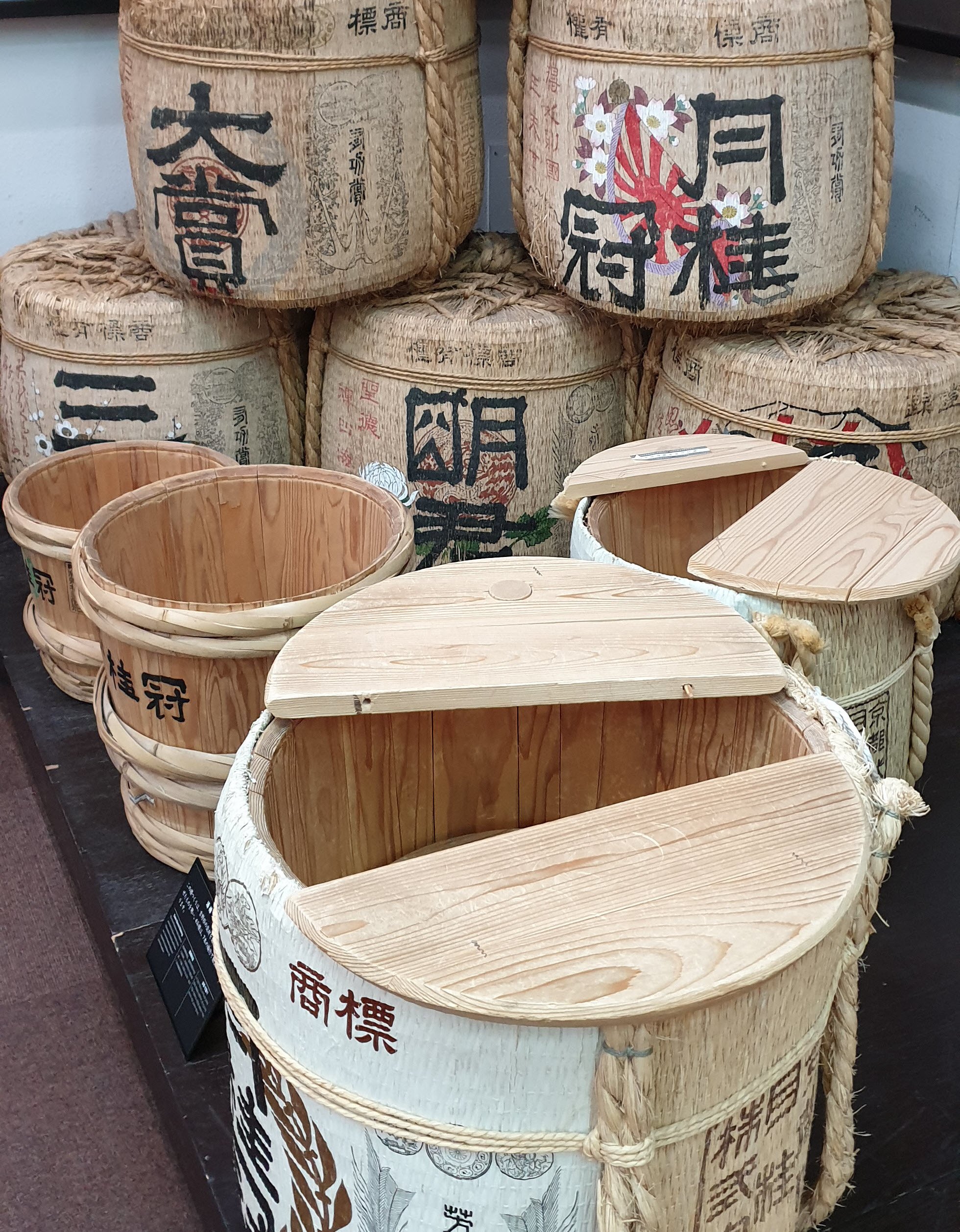 A collection of traditional Japanese sake barrels displayed in a stacked arrangement. The barrels are made of straw and wood, with various Japanese characters and decorative designs on their surfaces. Some of the barrels have their wooden lids open, revealing the interior.