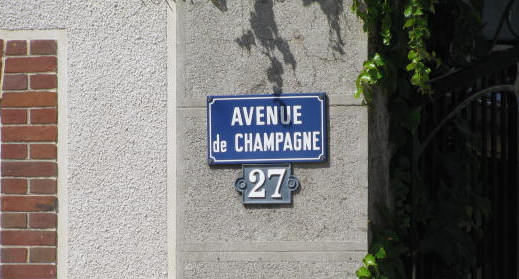 Blue street sign labelled 'Avenue de Champagne 27' on a grey wall with green leaves in the background.