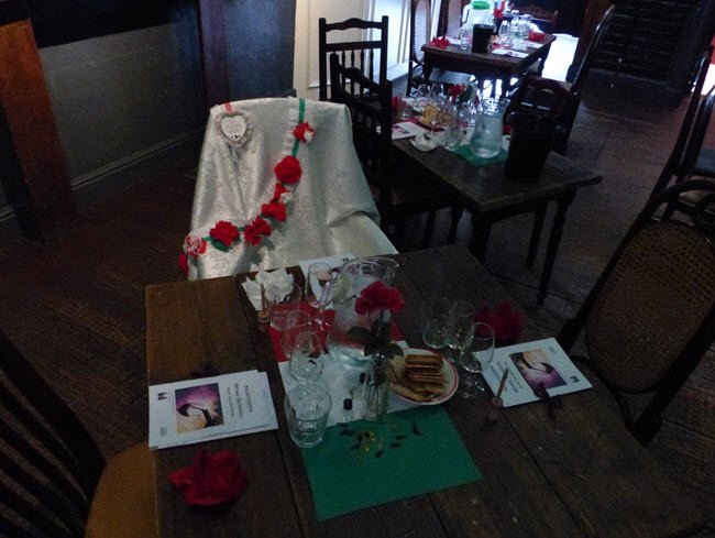 A dining table set up for a special occasion. The table is decorated with red flowers, a white cloth, and various glassware. There are booklets titled 'Northern Wine School' on the table, along with plates of food and a water pitcher.