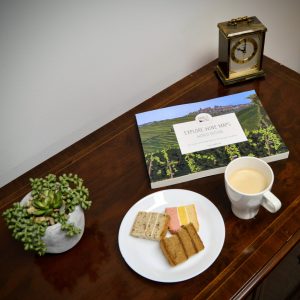 Explore Wine Maps" book on a wooden table with a plate of cakes, a coffee cup, a plant, and a vintage clock.