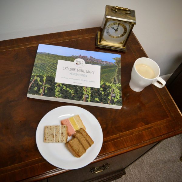 Explore Wine Maps" book on a wooden table with a plate of cakes, a coffee cup and a vintage clock.