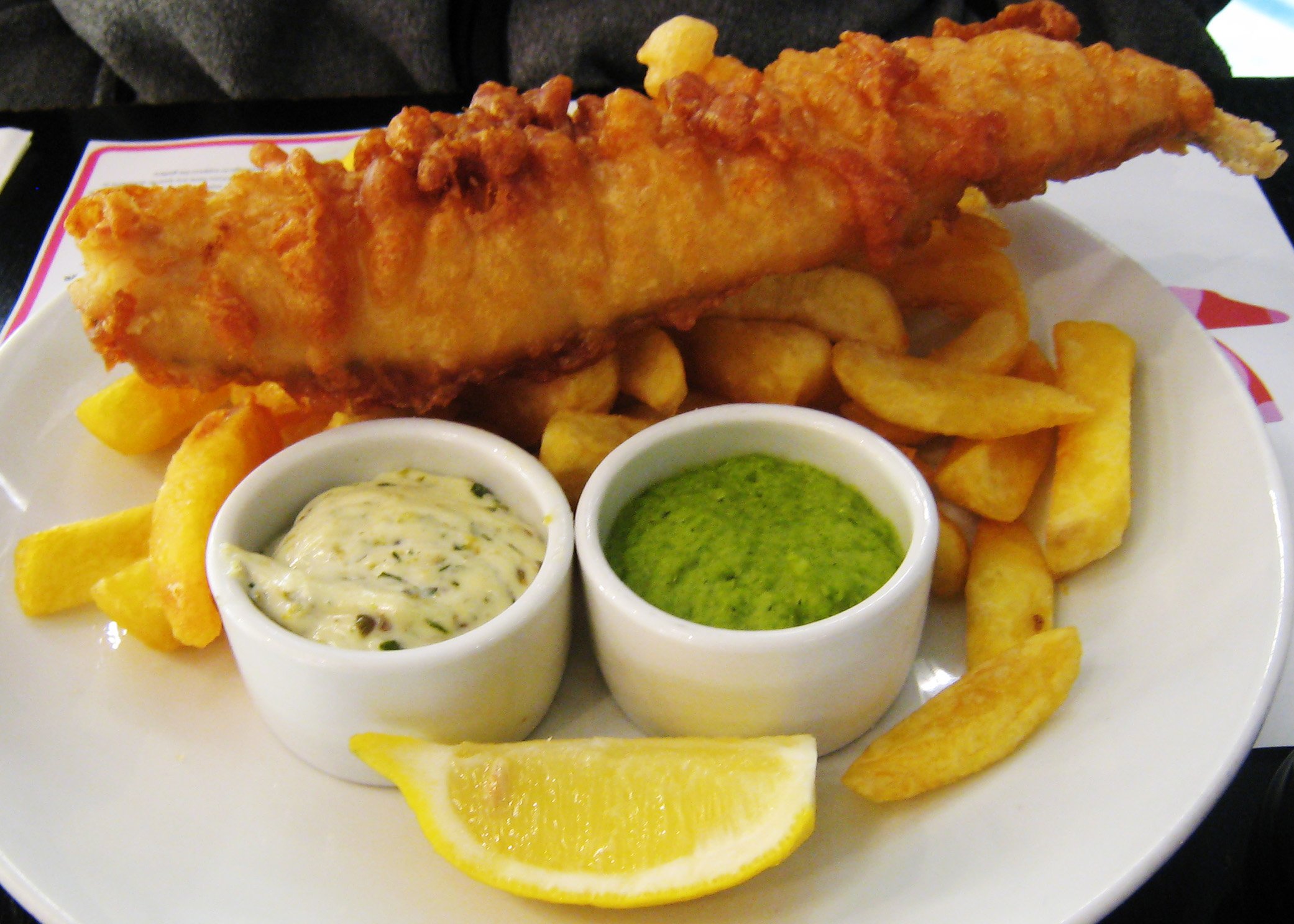 A plate of battered fish and chips, with a wedge of lemon, a small bowl of tartar sauce, and a small bowl of mushy peas on the side.