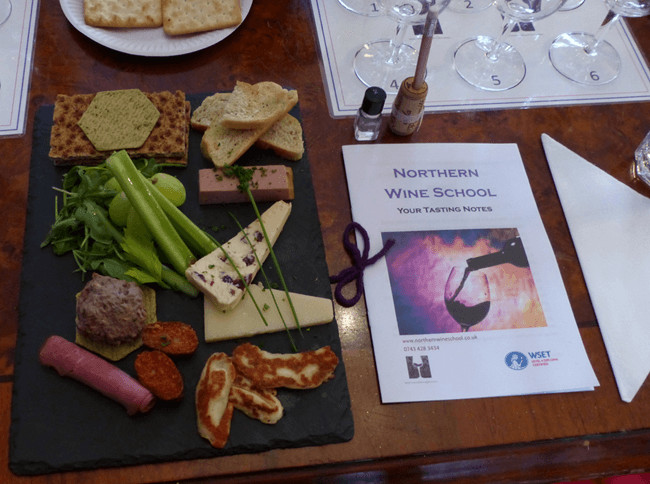 A charcuterie board with various cheeses, meats, vegetables, and crackers on a black slate, placed on a wooden table. Beside the board, there is a booklet titled 'Northern Wine School: Your Tasting Notes,' a white napkin, and a tasting setup with glasses and numbered mats. The table also has small bottles and a plate of additional crackers.