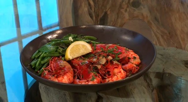 A black plate filled with cooked shrimp garnished with herbs, served with sautéed green beans and a lemon wedge, placed on a rustic wooden surface near a window.