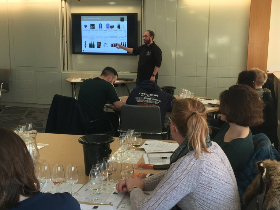 A group of people attending a wine tasting workshop, with a presenter pointing at a slide on a screen at the front of the room. The slide shows various types of wine bottles and glasses. Participants are seated at tables with wine glasses, notepads, and water pitchers in front of them.