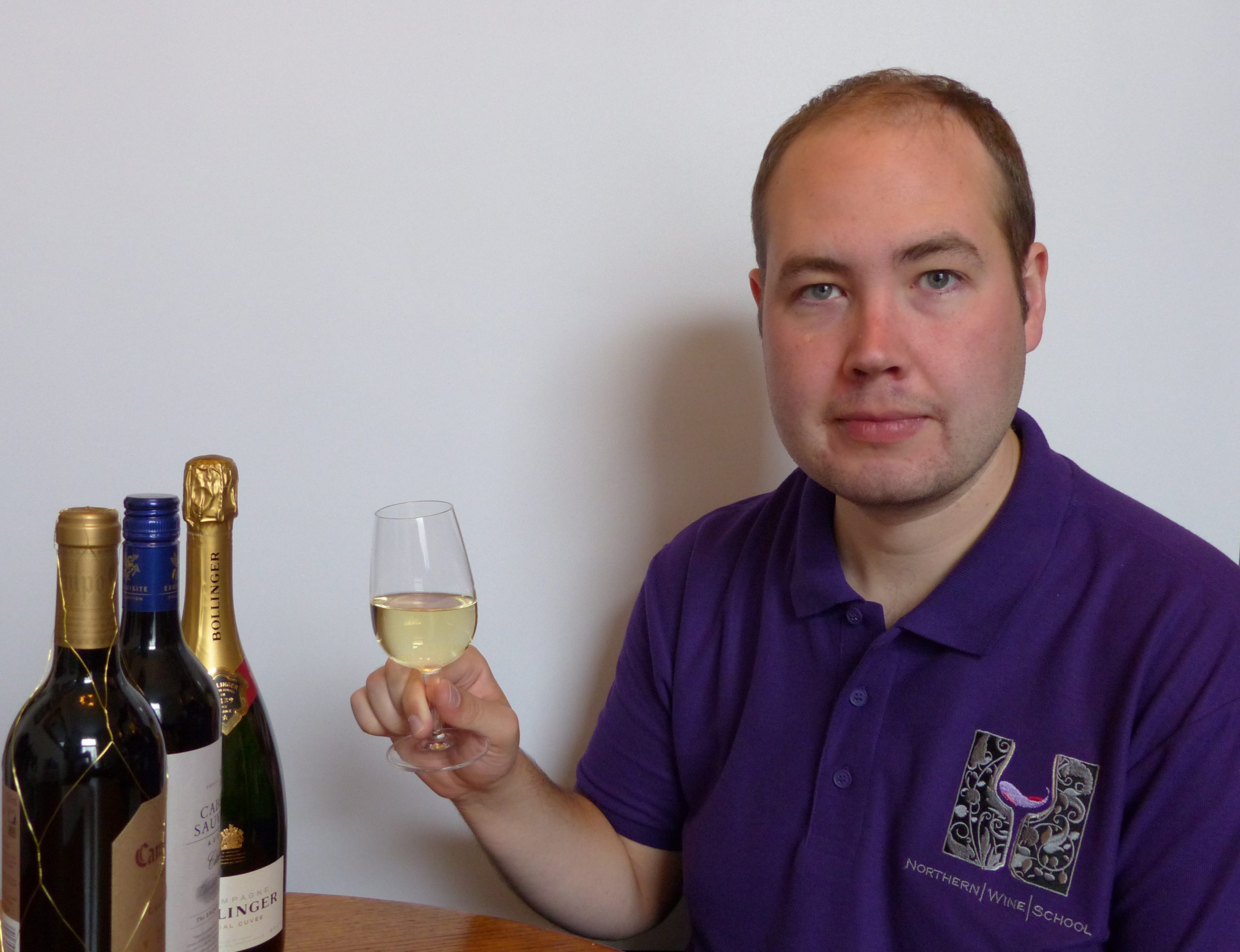 Man in a purple Northern Wine School shirt holding a glass of white wine, with wine bottles on the table beside him.