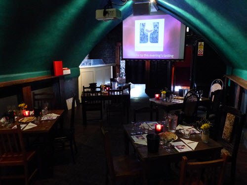 Dimly lit room set up for a wine tasting event with tables arranged with wine glasses and a projector screen displaying a welcome message.