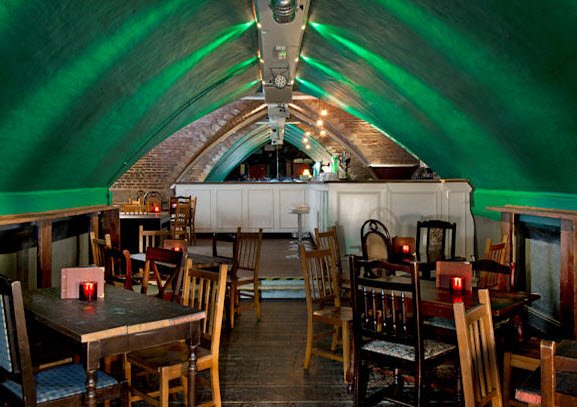 An underground bar with a green arched ceiling, wooden tables and chairs, and dim lighting.