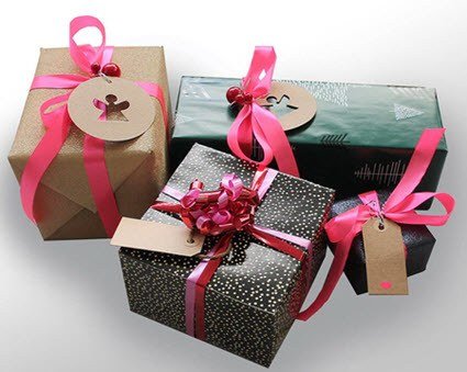 A group of three wrapped gifts of varying sizes, decorated with pink ribbons and bows. Each gift has a tag attached, with one tag featuring a cut-out gingerbread man. The wrapping papers are in shades of brown, black with gold dots, and green with white patterns.