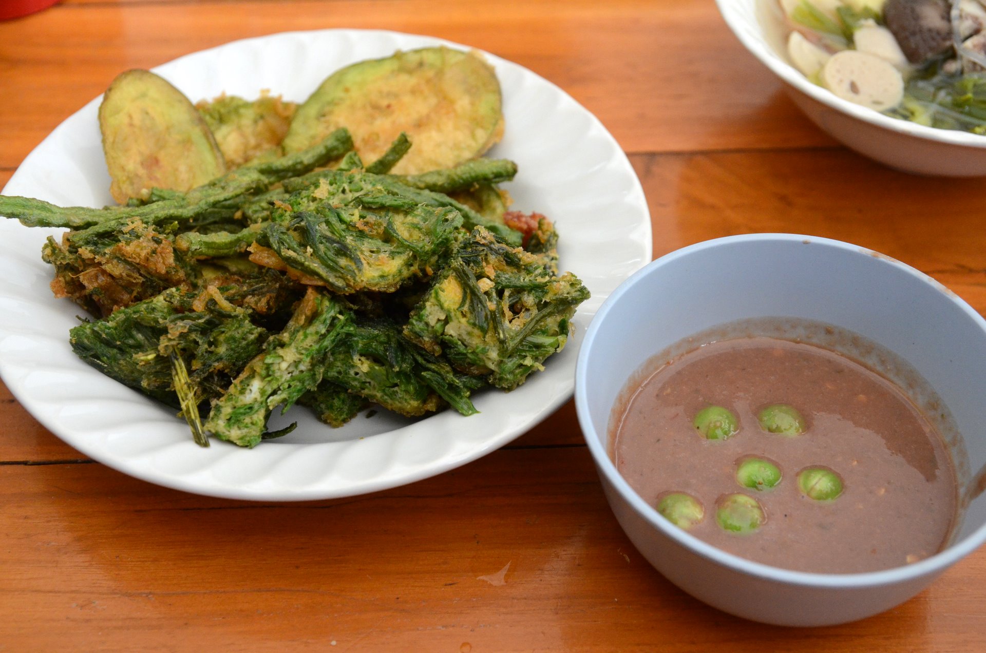 A plate of fried vegetables, including eggplant and green beans, served on a white plate. Beside the plate, there is a bowl of dipping sauce with green peas floating in it. The dishes are set on a wooden table.