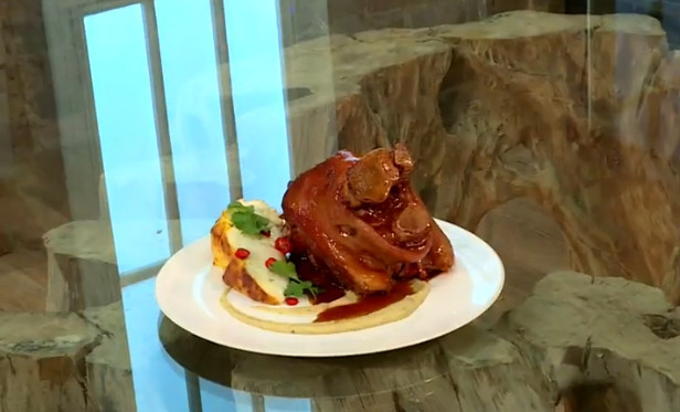 A plated dish featuring a succulent, glazed pork shank served with a side of mashed potatoes topped with herbs and pomegranate seeds. The dish is presented on a white plate, placed on a rustic wooden table near a window.