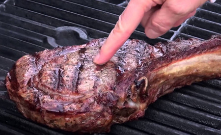 A close-up of a grilled steak on a barbecue grill with a person's finger pressing on it to check for doneness.