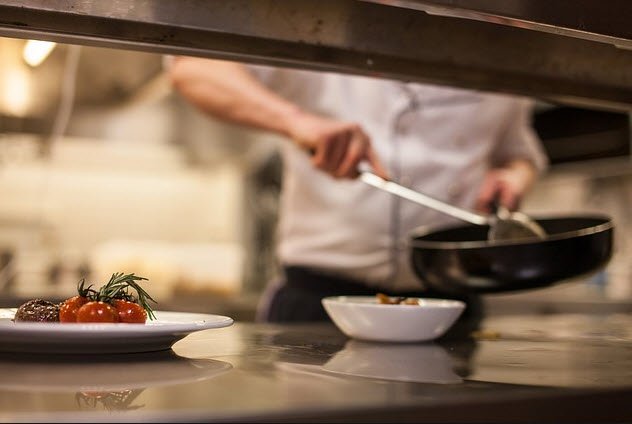 Chef in the background cooking with a pan, while plates of food are in the foreground, ready to be served.