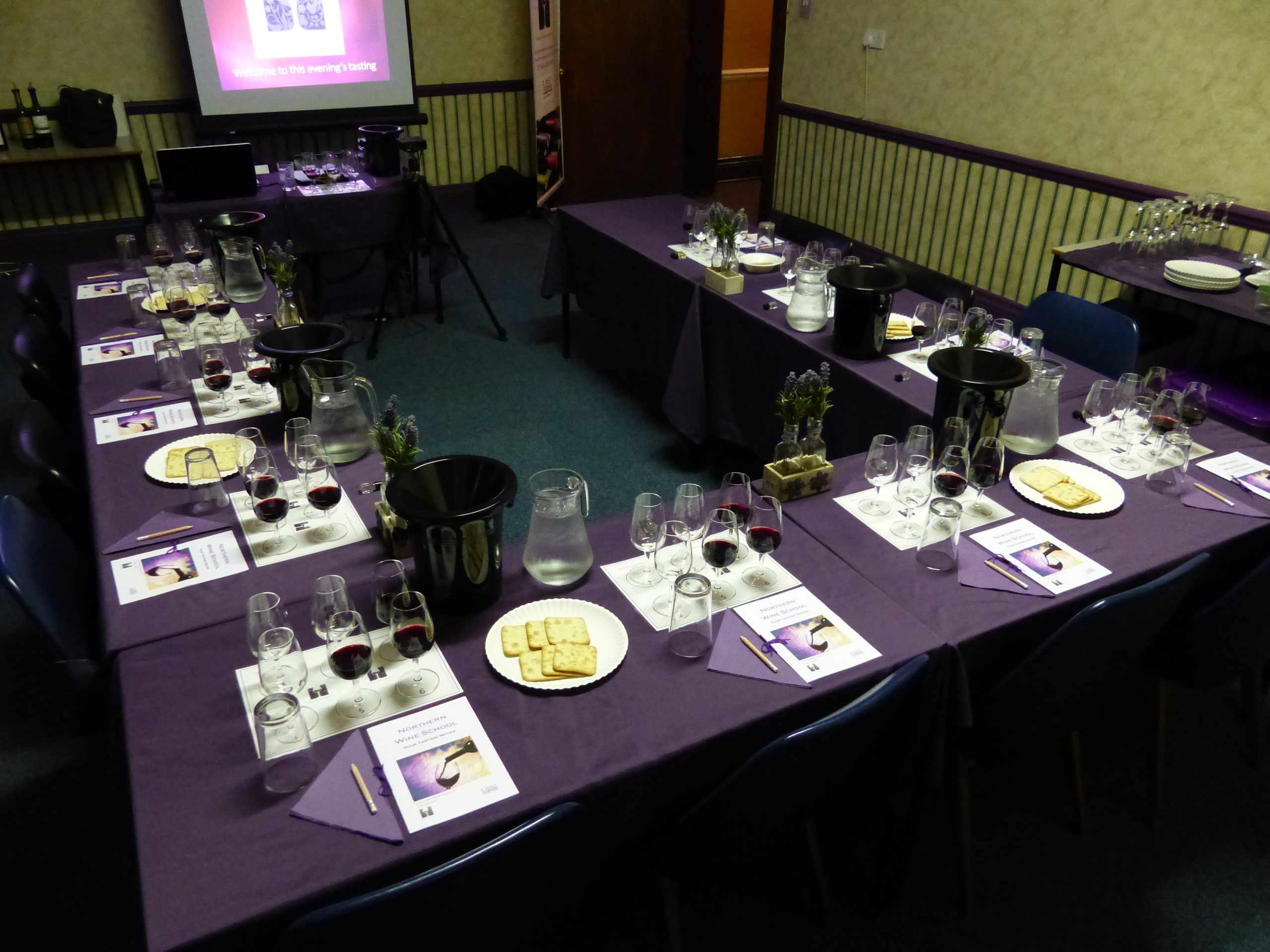 Room setup for a wine tasting event, featuring tables with glasses of red wine, crackers, and tasting notes.