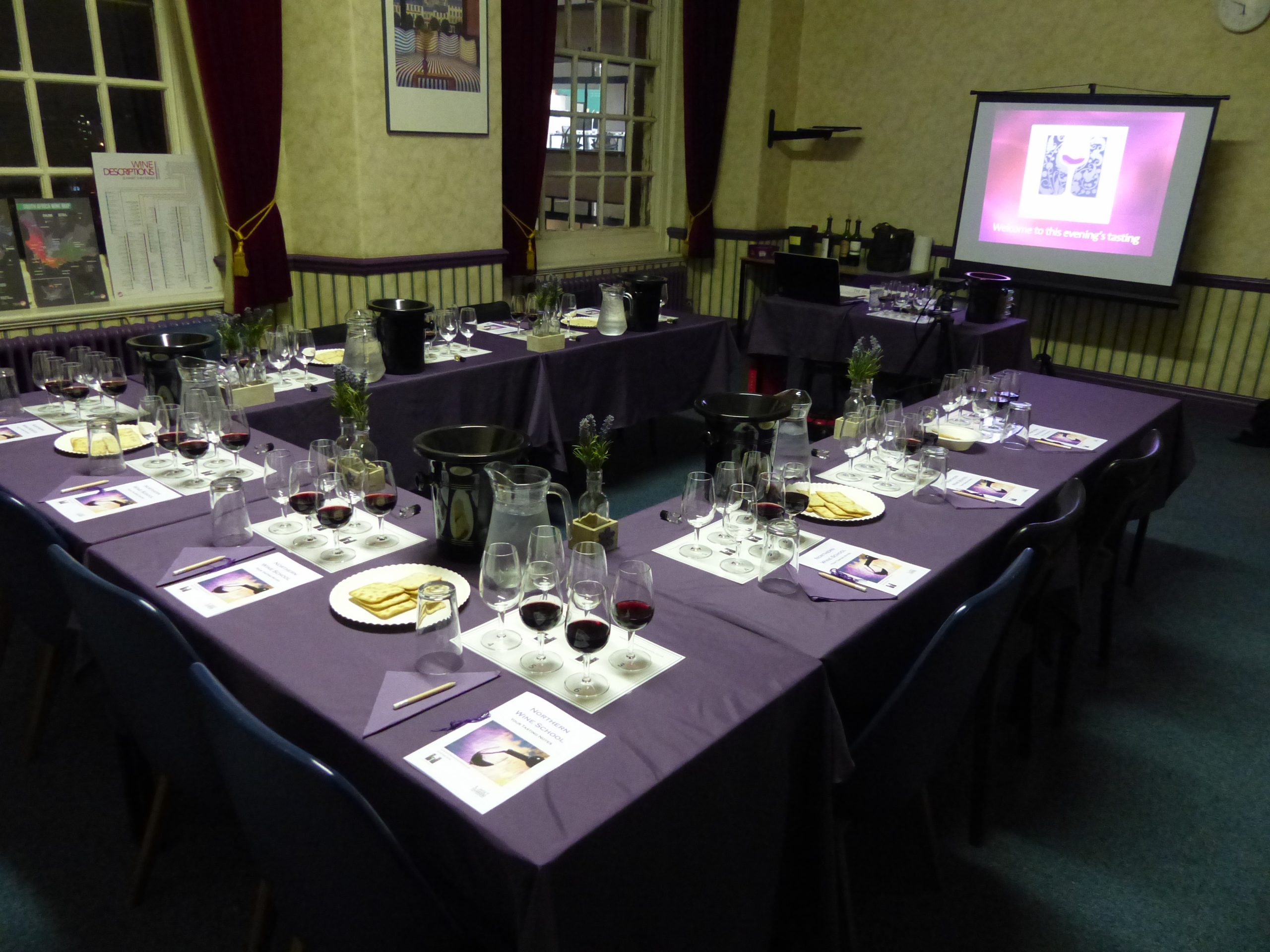 Tables set for a wine tasting event, featuring wine glasses, red wine, crackers, water jugs, and small flower arrangements.