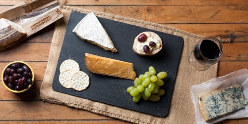 A cheese and grape platter on a black slate, with crackers and a glass of red wine on a wooden table.