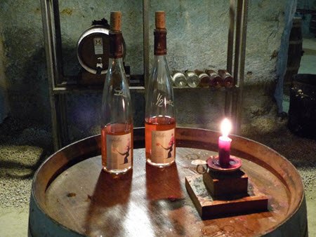 Two wine bottles and a lit candle on a barrel in a wine cellar.