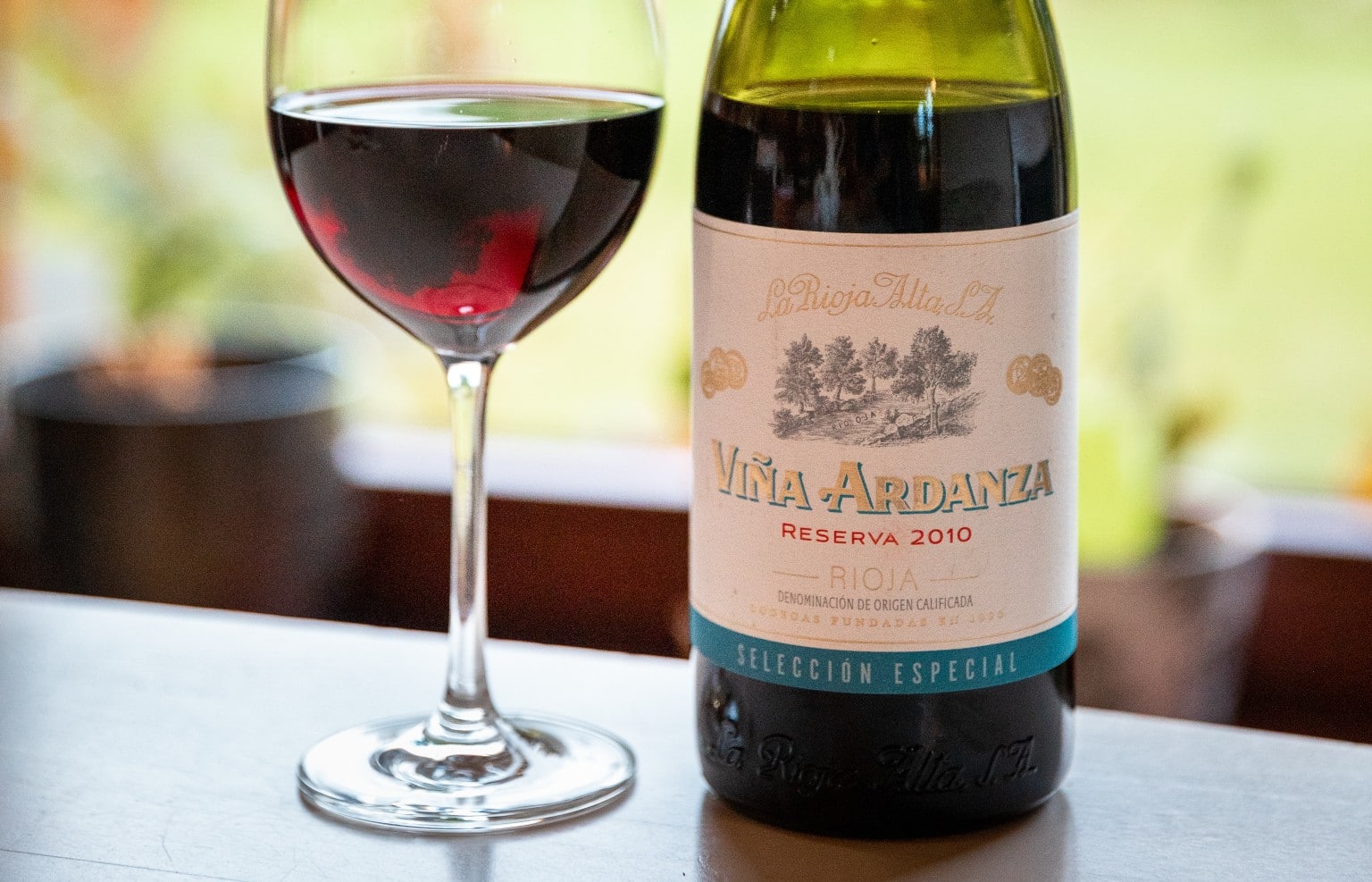 A bottle of Viña Ardanza Reserva 2010 Rioja wine next to a glass filled with red wine, set on a table with a blurred background of plants and soft lighting.