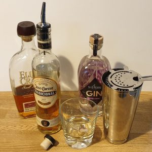 Whiskey being poured into a glass, with a gin bottle and cocktail strainer beside it on a wooden surface.