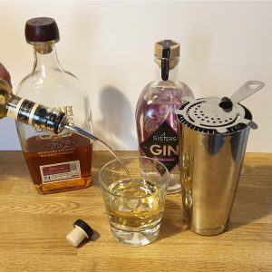 Whiskey being poured into a glass, with a gin bottle and cocktail strainer beside it on a wooden surface.