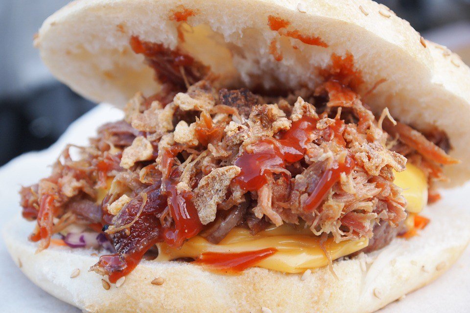 A close-up of a pulled pork sandwich with melted cheese, crispy onions, and a generous drizzle of barbecue sauce, served in a sesame seed bun.