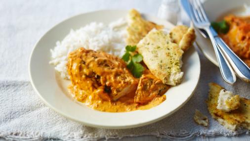 A plate of butter chicken served with white rice and garlic naan bread, garnished with a sprig of cilantro. The plate is set on a white cloth with a fork and knife placed beside it.