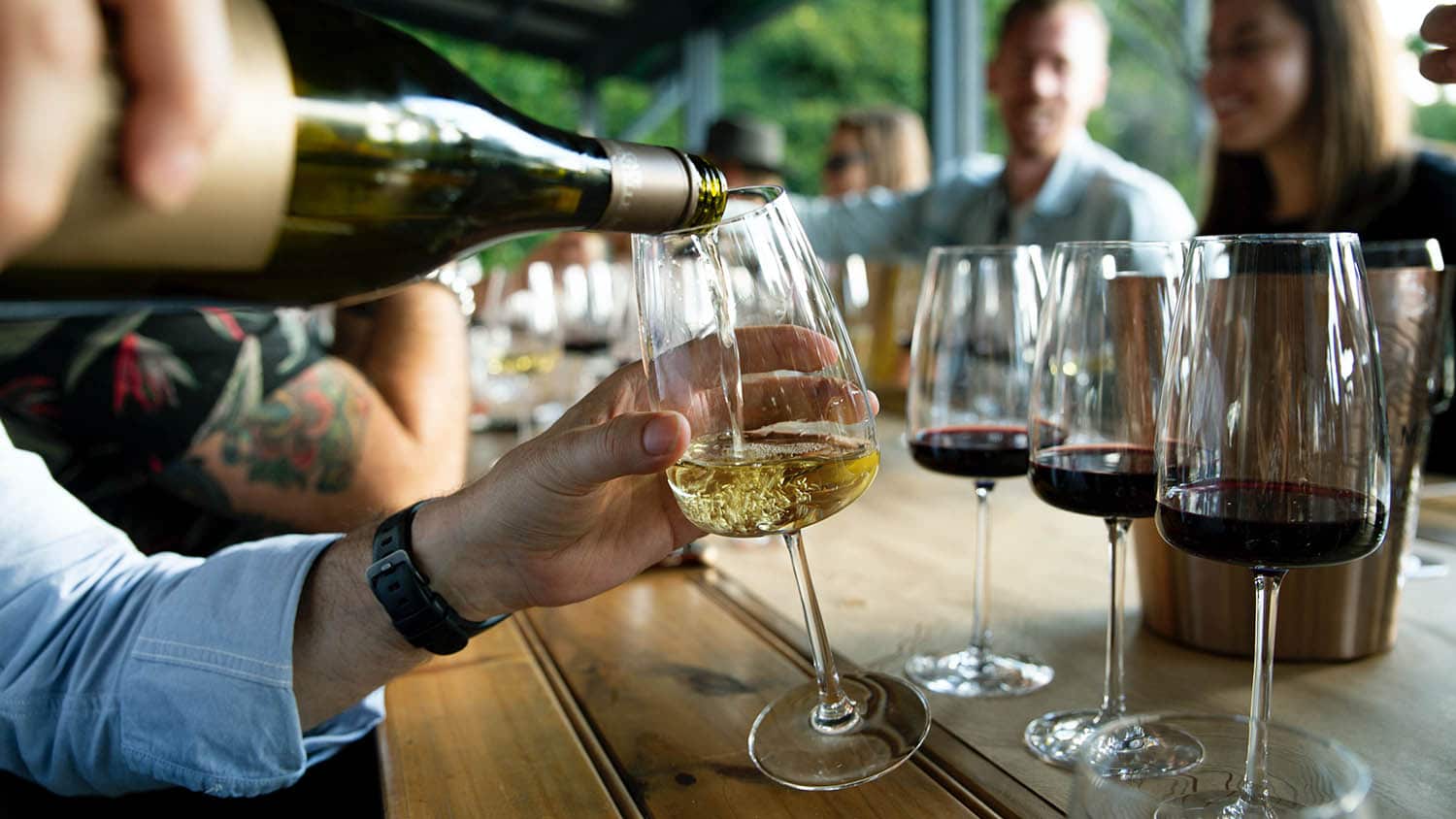 A group of people sitting around a wooden table, with one person pouring white wine into a glass. Several glasses of red wine are placed on the table.