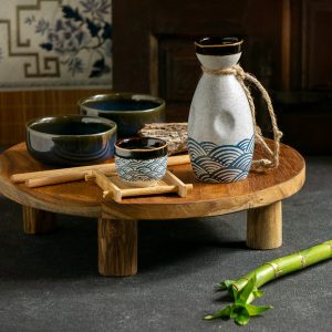 A small wooden table with two bowls, a cup, a decanter and some sticks on it. The is a piece of bamboo on the floor, next to the table.