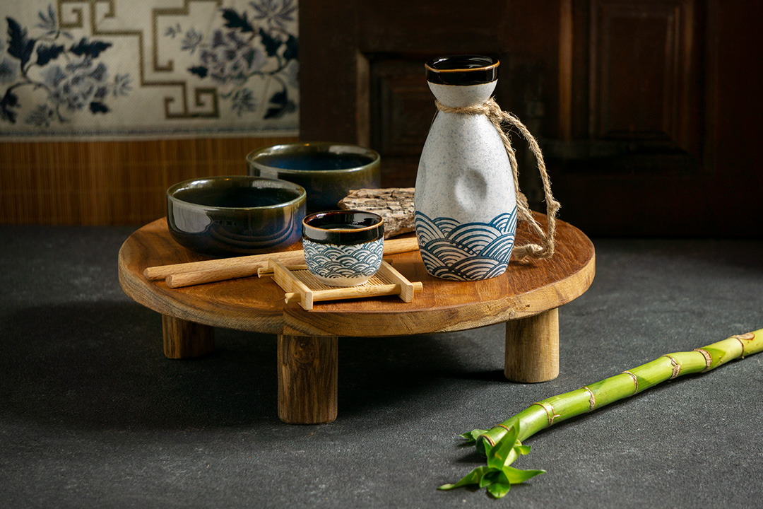 A small wooden table with two bowls, a cup, a decanter and some sticks on it. The is a piece of bamboo on the floor, next to the table.