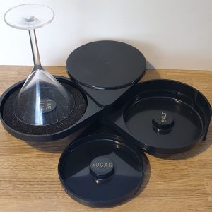 Black 3-tier rimmer set with sections for lime juice, salt, and sugar, holding an inverted martini glass.