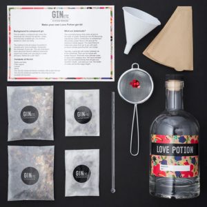 A "Love Potion" gin-making kit with various ingredients and tools, including a funnel, strainer, stirrer, and recipe card.