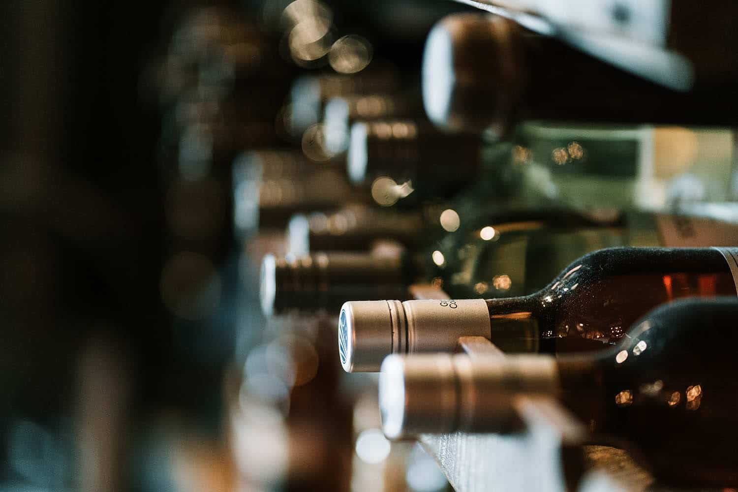 A close-up shot of wine bottles stored horizontally in a wine rack, focusing on the bottle caps. The background is blurred, creating a bokeh effect with warm lighting.