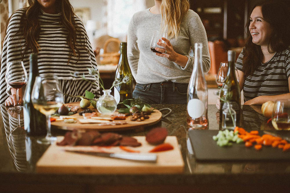 Three women enjoying a gathering around a kitchen island with various food items and wine bottles. The women are holding glasses of wine and appear to be engaged in conversation. The countertop is adorned with cutting boards containing sliced meats, cheeses, fruits, and vegetables. The background shows a cosy and well-lit kitchen.