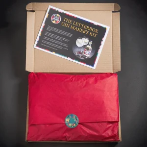 Closed gift box with a card on top labelled "The Letterbox Gin Maker's Kit." The box is wrapped in red tissue paper with a colourful sticker seal.