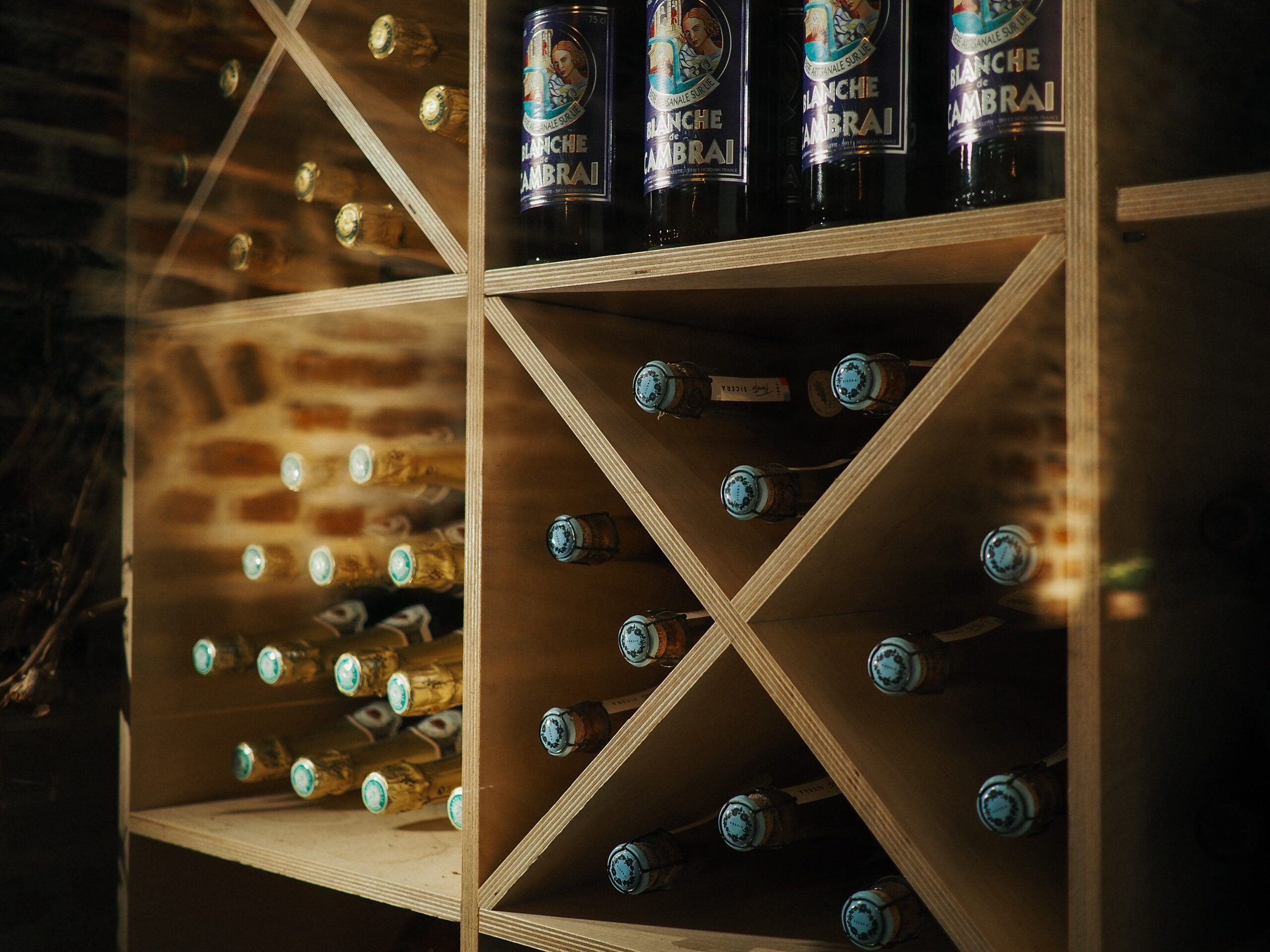 A wooden wine rack with several bottles of wine stored horizontally. The rack features a crisscross design, with some sections holding multiple bottles of wine with blue caps. The top section contains bottles of Blanche de Cambrai beer, each with a distinctive label.