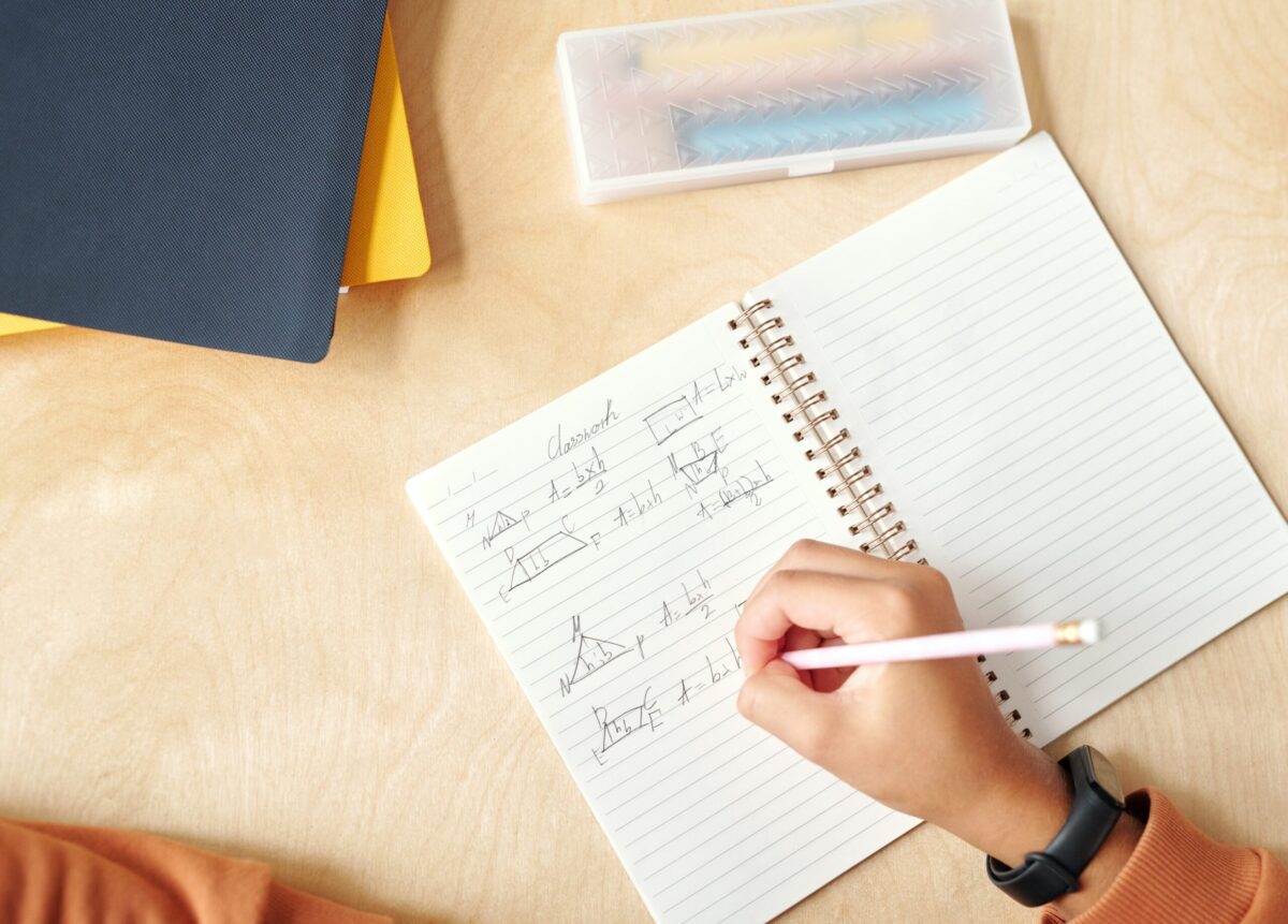 A person writing mathematical equations in a spiral notebook with a pencil. The notebook is on a wooden desk along with two closed books, one blue and one yellow, and a transparent pencil case containing coloured pens.