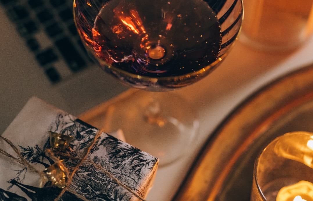 A close-up of a wine glass filled with red wine, positioned next to a small, wrapped gift tied with twine and gold bells. The background includes warm candlelight and part of a laptop keyboard