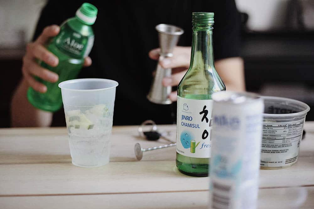 A person preparing a drink with a bottle of Jinro Chamisul soju and a green bottle of aloe beverage on a table. A plastic cup with ice and lime pieces, a jigger, and other beverage containers are also visible. The person is holding the aloe beverage bottle and the jigger while preparing the drink.