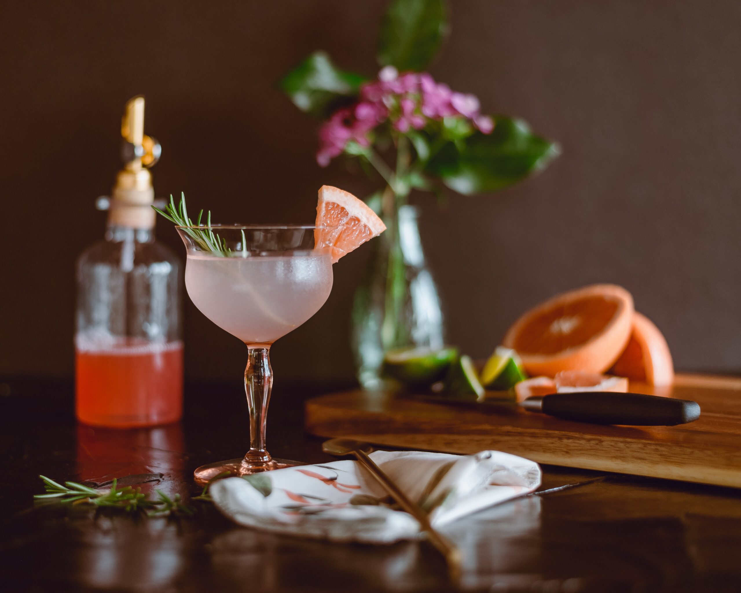 A delicate cocktail glass filled with a pink drink, garnished with a sprig of rosemary and a grapefruit slice, sits on a dark wooden table. In the background, there is a bottle with a red liquid, a cutting board with sliced grapefruit and lime, and a vase with pink flowers and green leaves.