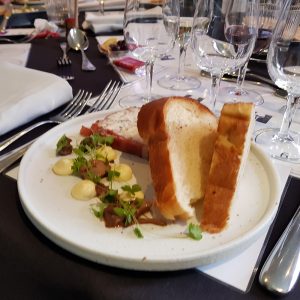 A plate with slices of bread, garnished with herbs and a creamy sauce, set on a formal dining table with multiple glasses and cutlery in the background.