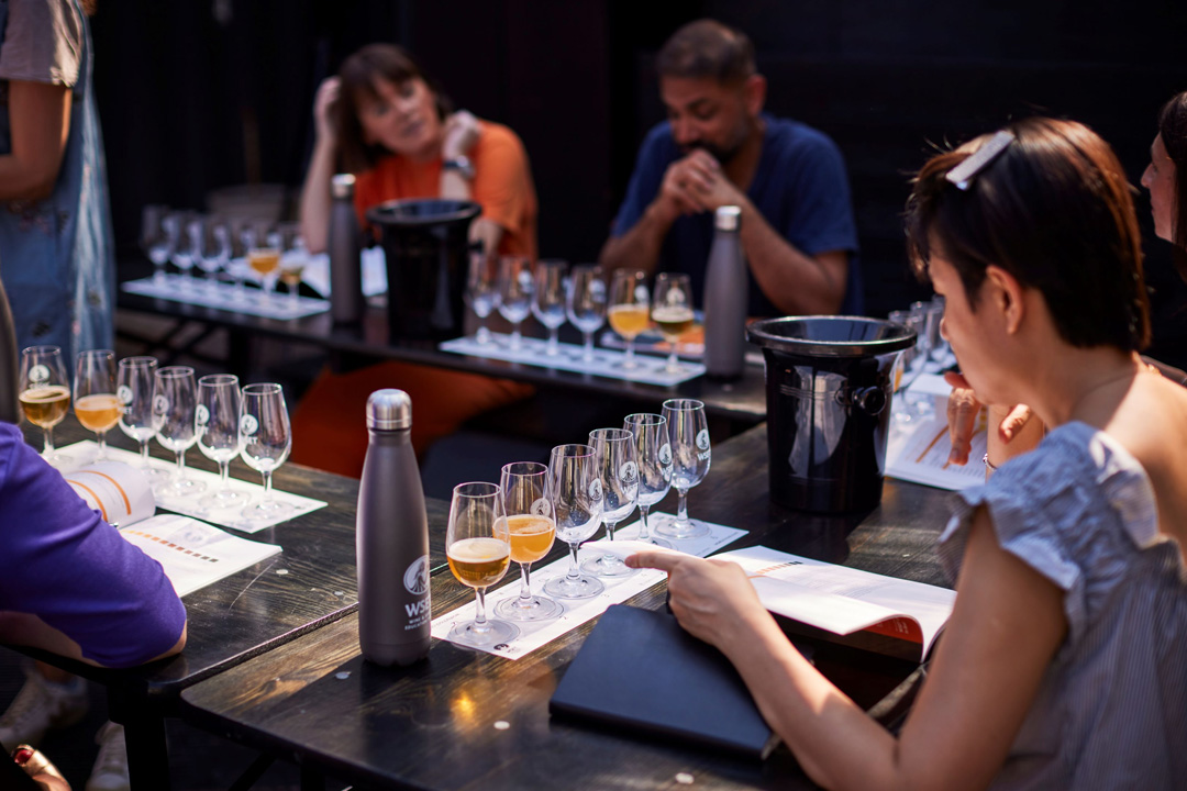 A group of people sitting at a u shaped table, looking at a book with samples of different beers in front of them.