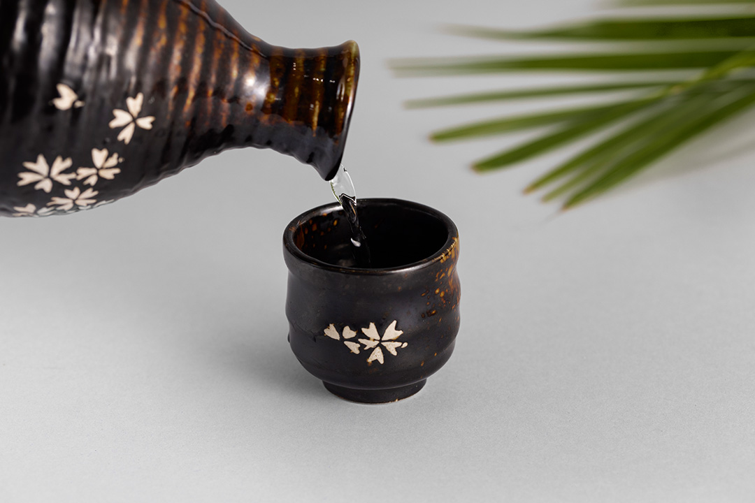 Pouring a clear liquid into a small, dark ceramic cup decorated with white floral patterns on a light background.