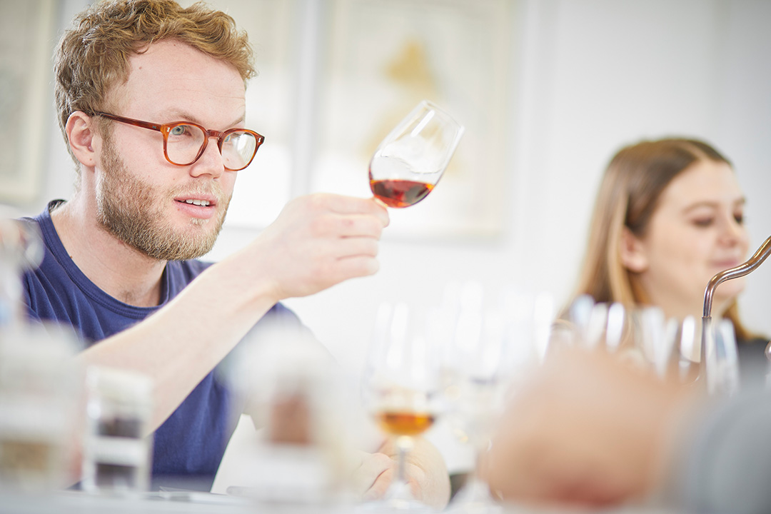 A man looking at a slightly tilted glass of spirits and a women in the background.