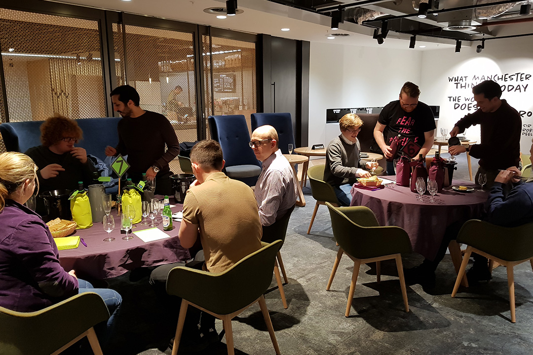 A group of people sat and stood around two tables tasting different wines and eating food.