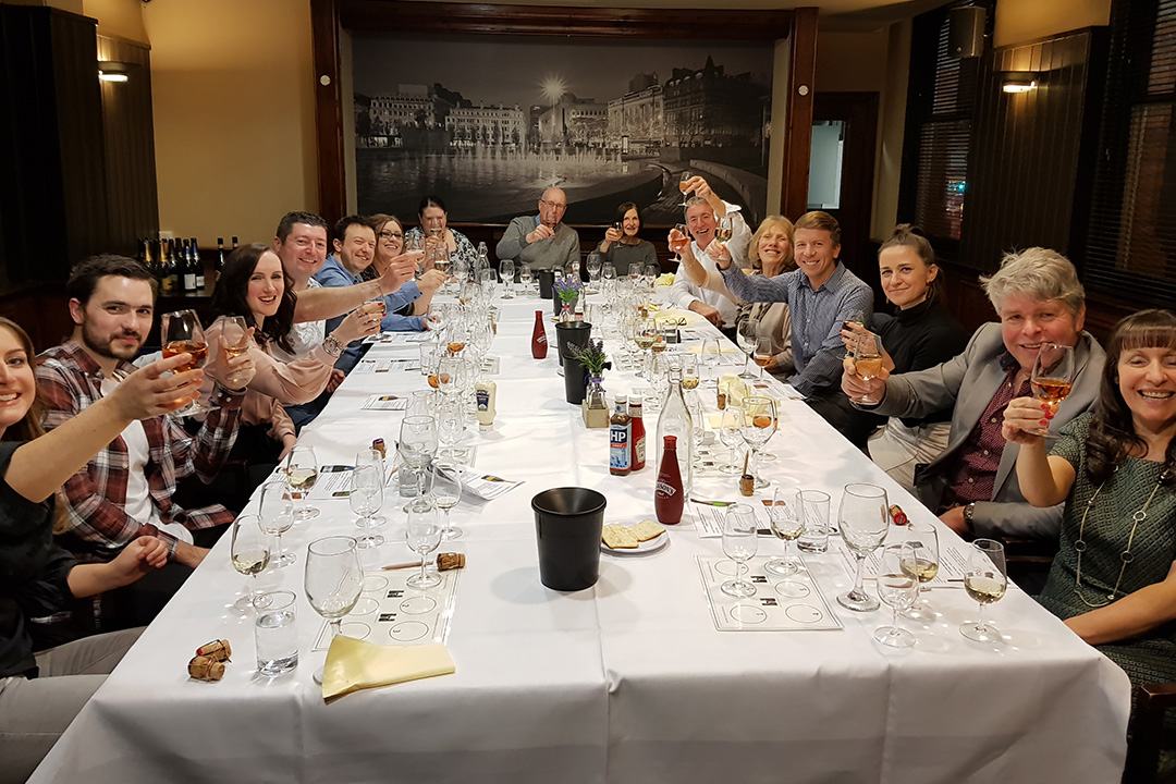 A group of people, sat round a large table, smiling at the camera and holding up their glasses.